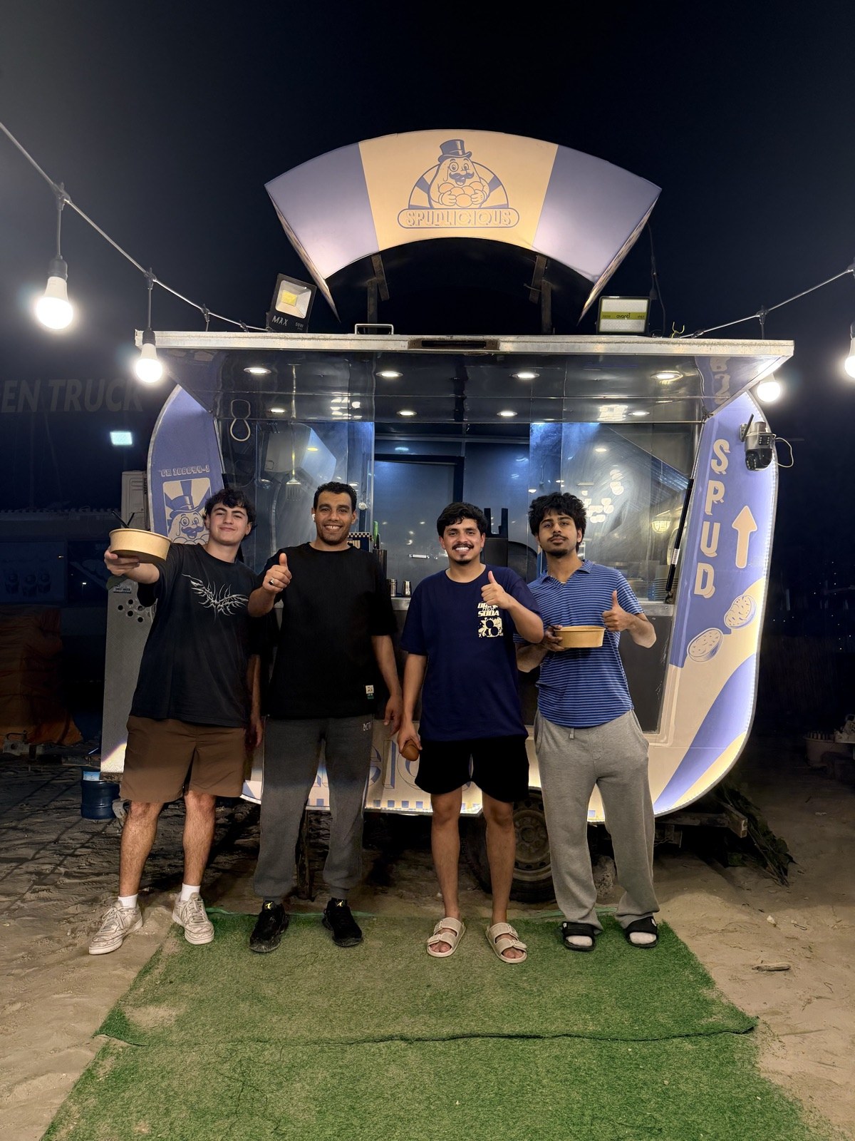The Spudlicious team and friends giving a thumbs-up with loaded potato bowls in front of the glowing truck